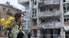 一名手捧花束的女子於 2025 年 5 月 25 日走過基輔謝甫琴科區的一棟高層住宅大樓。她的頭髮部分散落在額頭上。街道上,她身後是一棟遭俄羅斯無人機襲擊而嚴重受損的多層建築物。