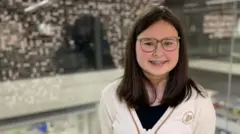 One teenage girl wit dark hair smile for di camera, wearing one white cardigan