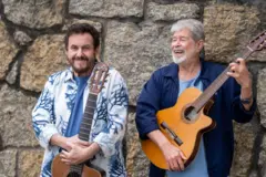 Antônio Carlos & Jocafi sorrindo em frente a muro de pedras, segurando violões