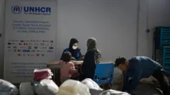 یک کارمند زن ملل متحد در تصویر واضح است که پشت ی میز نشسته و با یک خانم و دخترش صحبت میکند. یک مرد هم در گوشه راست تصویر دیده میشود.