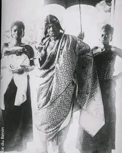 Photo archives du roi Behanzin portant un grand pagne autour de la taille avec une longue pipe entre les lèvres. Derrière lui se trouvent deux femmes dont l'une tient une parapluie pour protéger le roi.