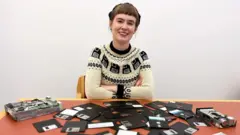 Seorang perempuan tersenyum dengan tangan terlipat duduk di depan disket-disket yang berserakan di atas meja. (Kredit: Perpustakaan Universitas Cambridge)