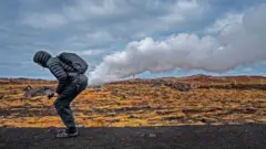Image d'illustration montrant un homme en doudoune courte à capuche, pantalon et chaussures de marche. Accroupi, il s'est positionné de telle sorte qu'un nuage semble provenir de son postérieur. Il se trouve sur une route bordant un paysage herbeux.