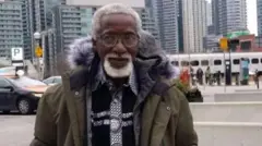 Baba Gebu wọ aṣọ òtútù, dúró sójú títí, gbogbo irun orí àti irungbọ̀n rẹ si funfun