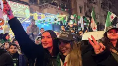 İki kadın kalabalık bir grubun arasında duruyor. Biri elini havaya kaldırmış durumda, diğeri fotoğrafçıya poz verirken eliyle zafer işareti yapıyor.