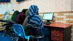 تصویر چهار دختر در یک صنف آموزش کمپیوتر. دختران در عقب میز و پشت شان به سمت دوربین نشسته اند. در مقابل شان یک کمپوتر دیده میشود. یک معلم زن هم در تصویر دیده می شود که به دختران رهنمایی میکند.