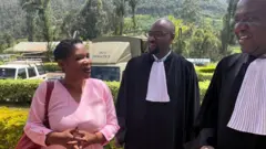 Béatrice Munyenyezi ahagararanye n'abunganizi be mu mategeko. Afite igikapu ku rutugu cy'ikigina, asobekeranyije intoki ndetse yambaye ikanzu y'iroza y'imfungwa. Abunganizi be bambaye amakanzu y'akazi y'umukara n'umweru mu gatuza. Bose uko ari batatu barimo kuvugana bamwenyura. Inyuma yabo hari ibiti n'imodoka zirimo n'itwara imfungwa.