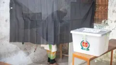 একটি পর্দার আড়ালে লুঙ্গি ও পাঞ্জাবি পরা একজন পুরুষ ভোট দিচ্ছেন, সামনে একটি বেঞ্চের ওপর ব্যালট জমা দেওয়ার একটি ভোট বাক্স