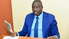 Faustin Ndikumana yambaye kositime n'ikaruvati yy'ubururu, n'ishati igomba kwera, afise igitabo mu kuboko kw'i buryo