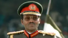 Muhammadu Buhari porte un uniforme militaire et des lunettes de soleil de type aviateur. Il porte une moustache.