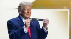 スーツに白シャツ、赤ネクタイ姿のトランプ氏が両手でくじをもって見せて笑顔を見せている