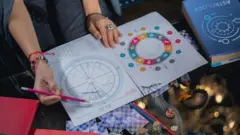 စားပွဲခုံတစ်ခုပေါ်မှာ ခံတံကိုကိုင်ထားပြီး နက္ခဗေဒ ဇယားပေါ်မှာ ညွှန်ပြနေတဲ့ အမျိုးသမီးတစ်ဦးရဲ့လက်ကို မြင်ရတာဖြစ်ပါတယ်။ စားပွဲပေါ်မှာတော့ ကတ်ဒ်တွေ ပြန့်ကြဲနေကြပါတယ်။