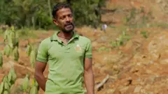 නායයෑමට ලක්වූ ස්ථානයක් පසුබිම්ව සිටින, කොළ පැහැති ටී-ෂර්ට් එකක් ඇඳ සිටින පුද්ගලයෙක්