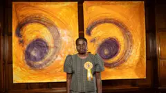 تصویر ننا کالو، هنرمند، که مقابل دو اثر طراحی خود با رنگهای زرد و بنفش ایستاده است