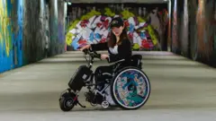 검은색 모자를 쓴 여성이 벽에 그래피티 벽화가 그려진 길 위에 휠체어를 타고 있다. 그가 탄 휠체어 바퀴에도 벽화와 비슷한 그래피티가 그려져 있다.