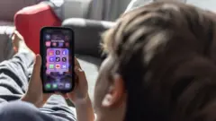 На фотографии 12-летний мальчик смотрит в свой телефон. В телефоне установлены приложения популярных соцсетей.