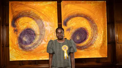 تصویر ننا کالو، هنرمند، که مقابل دو اثر طراحی خود با رنگهای زرد و بنفش ایستاده است