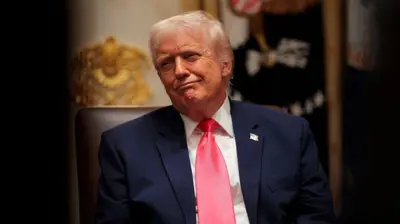 تصویری از دونالد ترامپ، رئیس جمهور امریکا که پشت میز خود نشسته و لبخنی معنیدار به لب دارد.