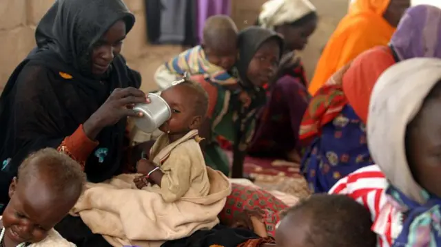 نساء سودانيات بعضهن برفقة أطفالهن، واحدة منهن تضع طفلاً على ظهرها وأخرى تطعم طفلها بوعاء يبدو أنه من الألمنيوم.