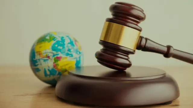 صورة تعبيرية تحتوي على المطرقة القضائية ذات اللون البني وخلفها كرة أرضية صغيرة على مكتب بلون بيج.