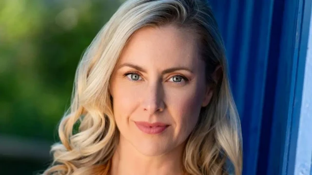 چهره زنی بلوند که به دوربین نگاه میکند پشت او یک پرده آبی رنگ دیده میشود