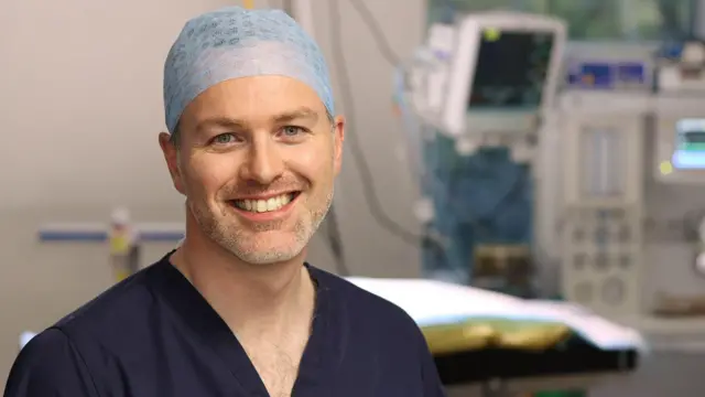 一名男子在手术室中微笑,头戴发网并穿着深蓝色医疗手术服