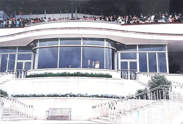 一张广角照片显示一栋大型白色建筑,带有玻璃窗,爱泼斯坦、安德鲁和麦克斯威尔站在其中一扇窗前,他们上方是人群