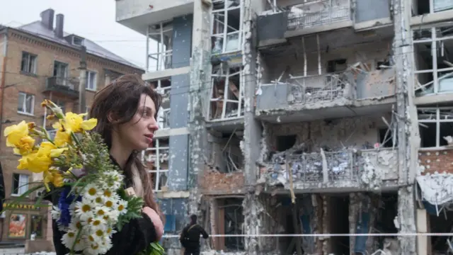 زنی با یک دستهگل از مقابل یک ساختمان بلند مسکونی در منطقه شوچنکیوسکی کییف، در ۲۵ مه ۲۰۲۵ عبور میکند. بخشی از موهای او روی پیشانیاش ریخته است. در پسزمینه، ساختمان چندطبقهای دیده میشود که در حمله پهپادی روسیه بهشدت آسیب دیده است.