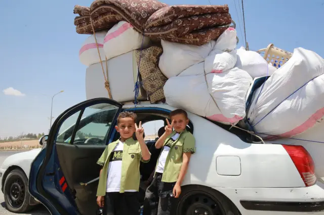 Two boys gesture as a Syrian refugee family moves in a car loaded with belongings