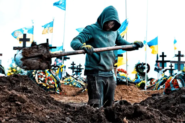 حفر قبر در گورستانی در اودسا در ۱۶ مارس ۲۰۲۴. حدود ده پرچم اوکراین بر روی میلههای سفید در پسزمینه به اهتزاز درآمده است. مردی که در پیشزمینه دیده میشود یک لباس کلاهدار بر تن و دستکش زرد به دست دارد.