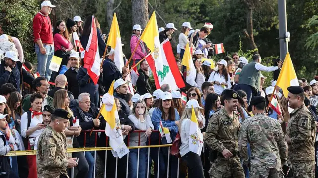 حشود من اللبنانيين من رجال ونساء يقفون على جانب الطريق وخلف حواجز وهم يحملون أعلام الفاتيكان والأعلام اللبنانية للترحيب ببابا الفاتيكان الزائر الباب ليو الرابع عشر.