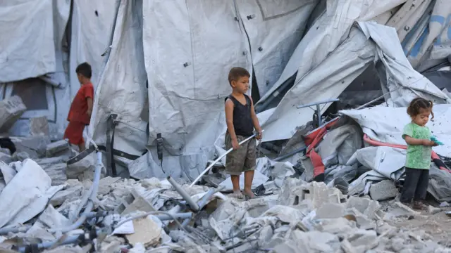سه کودک، دو پسر و یک دختر در شهر غزه در میان آوار ایستادهاند و چادرهایی پشت سرشان قرار دارد. پسر در مرکز تصویر یک لوله پلاستیکی در دست دارد. دختر سمت راست با یک اسباببازی پلاستیکی بازی میکند، ۹ سپتامبر ۲۰۲۵
