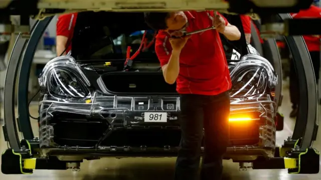 在德国的一家工厂里,一名身穿红色T恤的男子正在用工具修理汽车底部,他身后停着一辆正在组装的汽车。