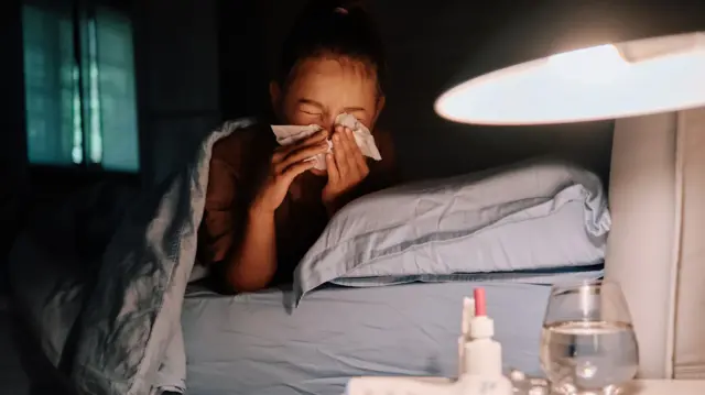 A person tucked under a duvet in a dark room blows their nose into a tissue. A glass of water, thermometer and medicine sit on a side table.