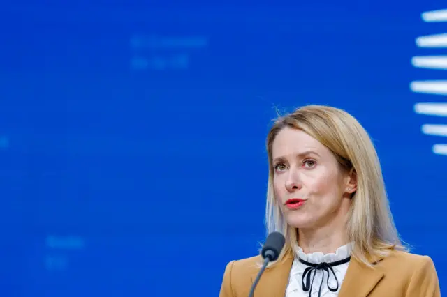 کایا کالاس، مسئول سیاست خارجی اتحادیه اروپا، با ژاکت بژ و بلوز سفید با حاشیه دانتل مشکی، در برابر پسزمینه آبی روشن با عناصر گرافیکی سفید، درون میکروفون صحبت میکند.