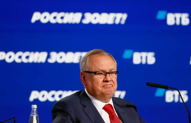 آندری کوستین، رئیس هیئت مدیره بانک دولتی روسی ویتیبی، با عینک و کراوات قرمز در برابر پسزمینه آبی روشن با نوشتههای روسی و لوگوی ویتیبی نشسته است.