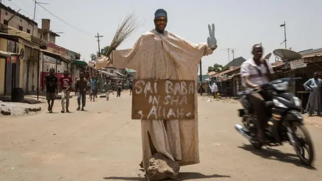 Au milieu d'une route poussiéreuse se dresse une statue de Buhari, portant l'inscription « Sai Baba Inshallah ». Une moto passe en trombe.
