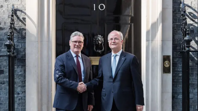 کییر استارمر و محمد مصطفی روی پلههای خیابان داونینگ شماره ۱۰ ایستادهاند و دستهایشان را محکم به هم میفشردند. در مشکی و براق اقامتگاه با شماره ۱۰ درست پشت سر آنهاست. هر دو مرد کت و شلوار سورمهای پوشیدهاند. استارمر کراوات قرمز تیره و مصطفی کراوات آبی روشن دارد. نور خورشید روی در و چارچوبهای سفید در میتابد