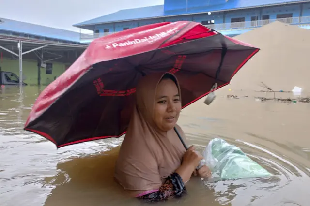 زنی که چتری بهدست دارد و درحالیکه بکسی را به محل امنی حمل می کند در میان آبهای سیل قهوهای رنگ اندونزیا دیده میشود که به صورت سینهخیز پیش میرود.