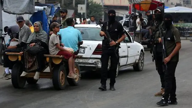 سه نیروی حماس که صورتهایشان را پوشانده شده، در خیابانی در غزه گشت میزنند. یک ماشین سفید، گاری زرد کوچکی را میکشد که یک زن، چند مرد و یک پسر در آن نشسته و پاهایشان بیرون آویزان است
