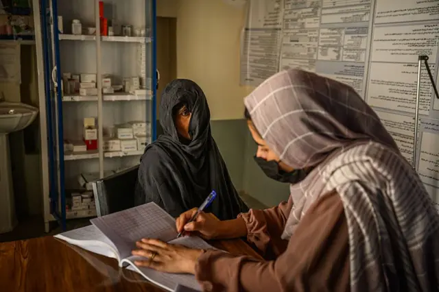 یوه ډاکټره چې مخ یې په تور ماسک پوښلی، برګ ټېکری یې پر سر او نسواري کالي یې اغوستي، په کتابچه کې یو څه لیکي او تر څنګ یې یوه ناروغه ښځه ناسته ده چې مخ یې نیمه پټ دی.