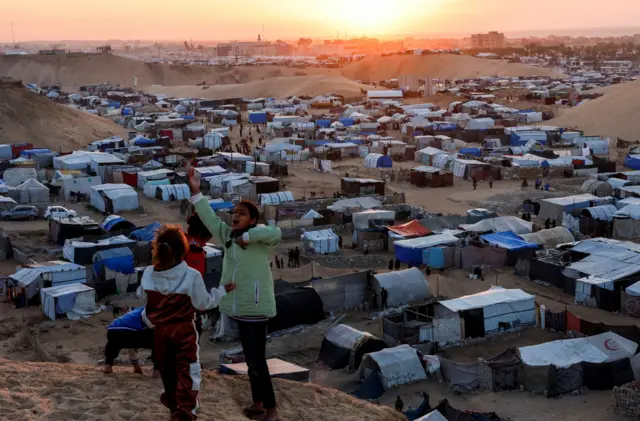 دو دختر کودک فلسطینی بر فراز تپهای مشرف به اردوگاه چادری در خان یونس، نوار غزه، در حال صحبت با هم یکی از آنها دستش را بالا برده است. پشت سر آنها چادرهایی قرار دارد که خورشید در افق در حال غروب است. ۲۹ دسامبر ۲۰۲۴ عکس از محمد سالم / رویترز