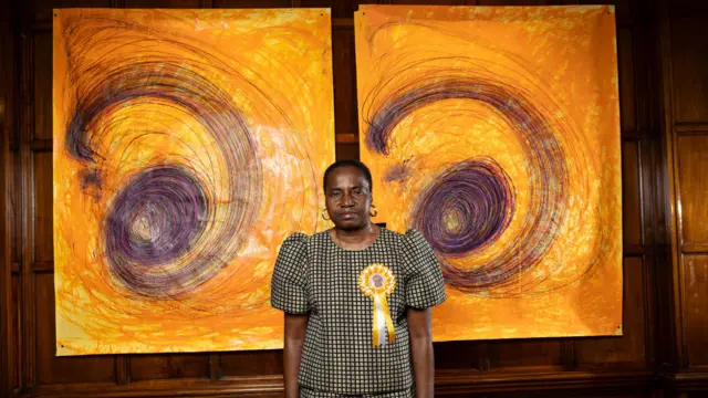 تصویر ننا کالو، هنرمند، که مقابل دو اثر طراحی خود با رنگهای زرد و بنفش ایستاده است