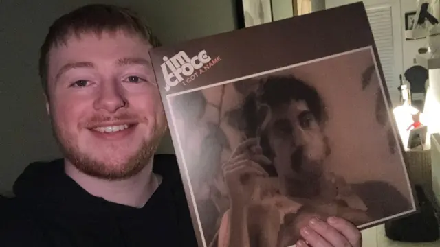 索尔穿着黑色连帽外套,手中拿着两张黑胶唱片。背景是一面绿色墙壁,上面挂有装裱的艺术作品和一面圆形镜子