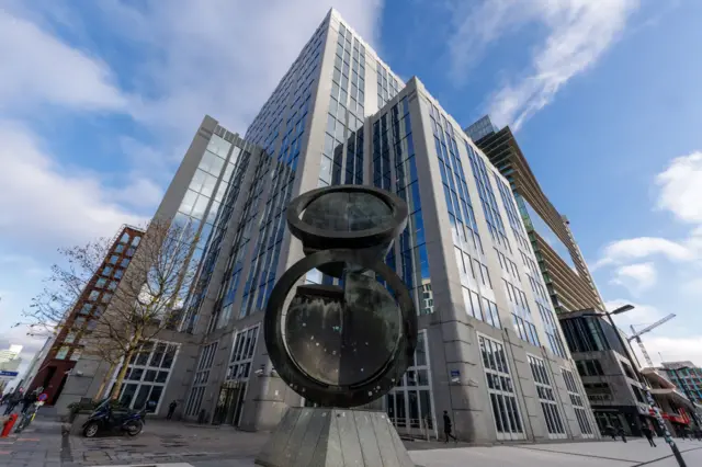 ساختمان بلند و مدرن یوروکلیر با پنجرههای شیشهای بازتابنده، از زاویه پایین عکاسی شده؛ مجسمه فلزی دایرهای بزرگی در پیشزمینه دیده میشود و آسمان آبی با چند تکه ابر در بالای آن قرار دارد.