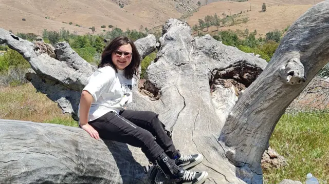 فتاة مراهقة جالسة على جذع شجرة ساقط ذي لحاء باهت بفعل الشمس.