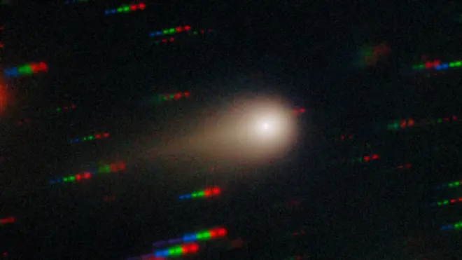 彗星“3I/Atlas”呈现为明亮核心,周围环绕朦胧的彗发,左侧延伸出彗尾;背景中可见蓝、绿、红色的光点条纹,代表相对于彗星移动的恒星。
