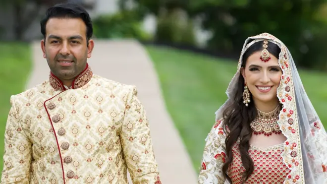 Sammi and Safi dressed in their wedding atire posing for a photo and holding hands - Sammi is wearing a traditional white and red bridal Afghan dress and wearing a scarf on her head, while Safi is wearing a golden traditional groom's outfit called a Sherwani.