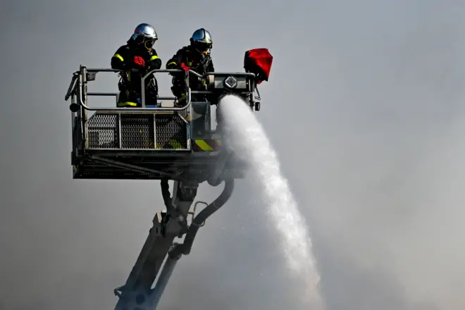 Two firefighters on a platform with a water hose and a ladder with rescue basket.