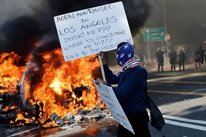 يقف الناس في الشوارع بجانب سيارات وايمو المحترقة في لوس أنجلوس، كاليفورنيا. يبرز في الصورة ألسنة نيران مشتعلة وأحد المحتجين الملثمين يحمل يافطة كتب عليها ولدت وأعيش في لوس أنجلوس، تريد ترحيلي؟؟؟